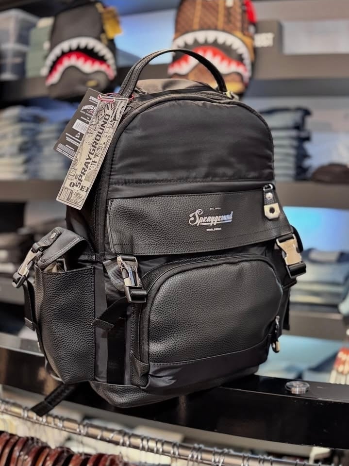 Black textured backpack with multiple pockets and handles displayed on a counter with branded merchandise in the background