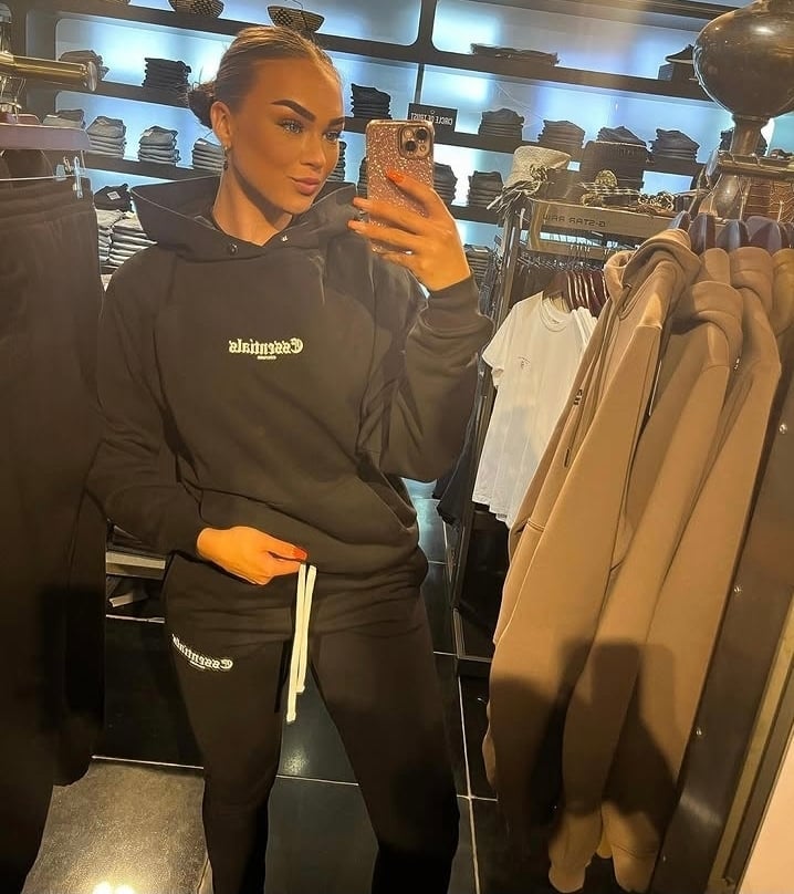 Woman taking a mirror selfie in a retail clothing store, wearing a black hoodie and holding a phone to her face