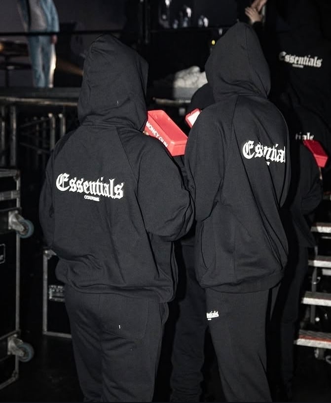 Two people wearing black Essentials hoodies viewed from behind at a nighttime event or venue