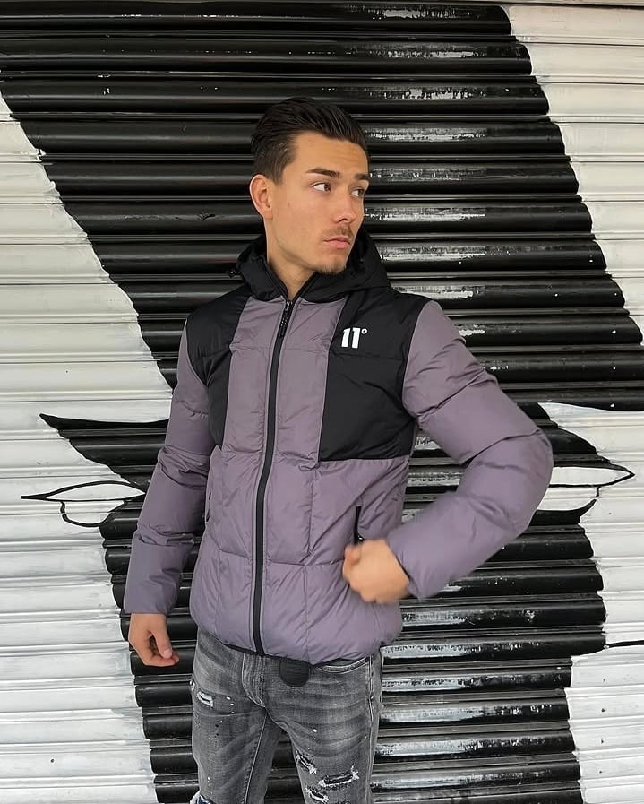 Young man wearing purple and black puffer jacket and distressed jeans posing against a corrugated metal wall