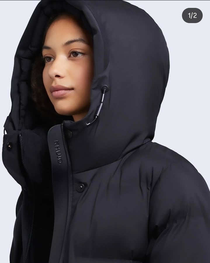 Person wearing a black hooded puffer jacket with hood up, looking to the side against a light gray background